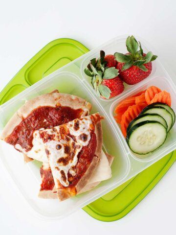 sliced pita pizza in a lunch container with strawberries, cucumber, and carrots.