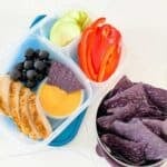 lunchbox filled with sliced chicken, fruit and sliced veggies, purple chips and cheese dip