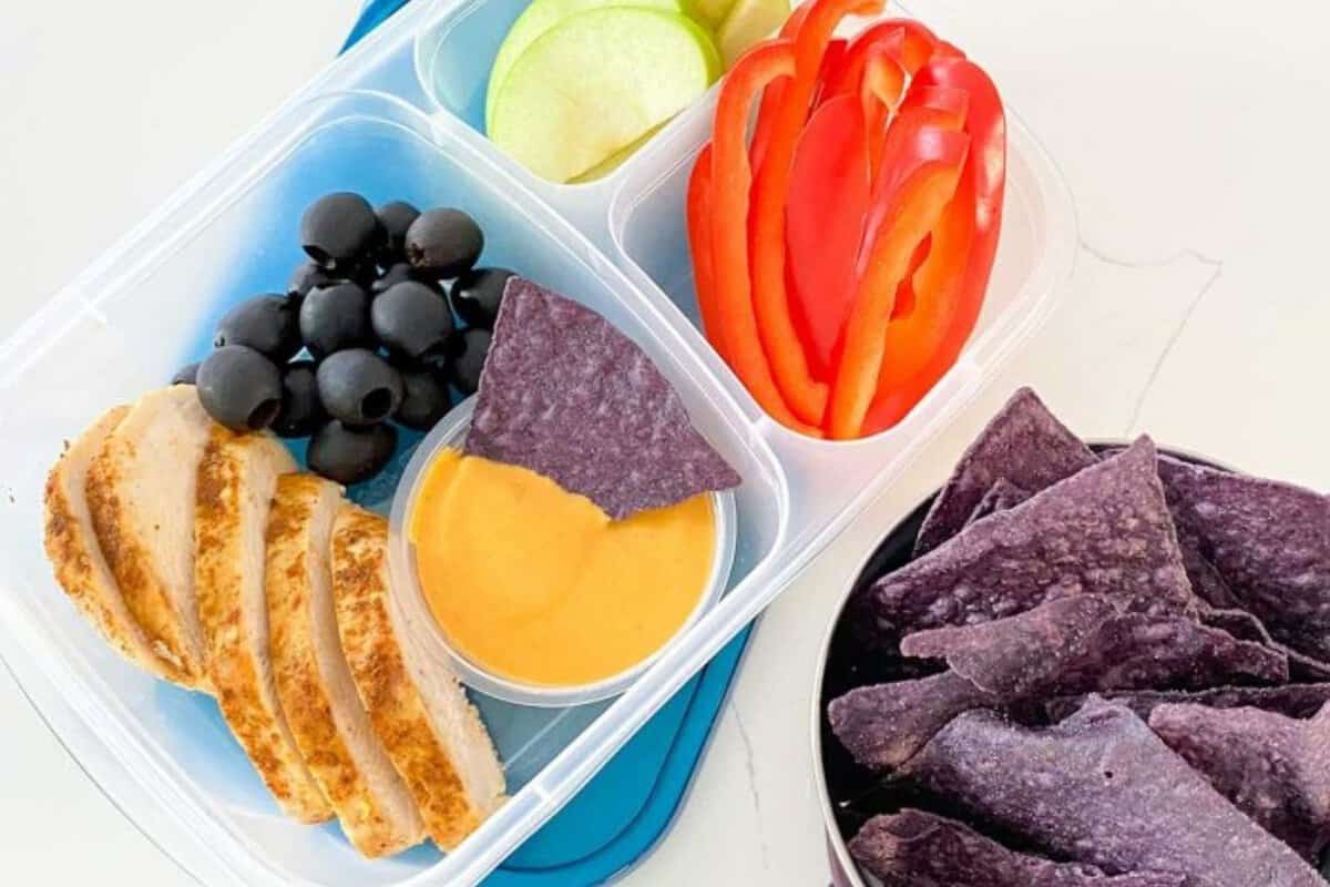 lunchbox filled with sliced chicken, fruit and sliced veggies, purple chips and cheese dip