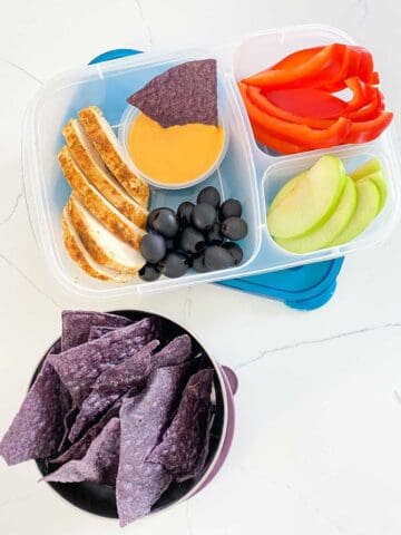 lunchbox filled with sliced chicken, fruit and sliced veggies, purple chips and cheese dip