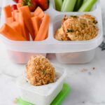 a chicken nugget being dipped into a sauce cup of Ranch dressing with a lunch container filled with chicken nuggets, veggies, and strawberries, in the background.
