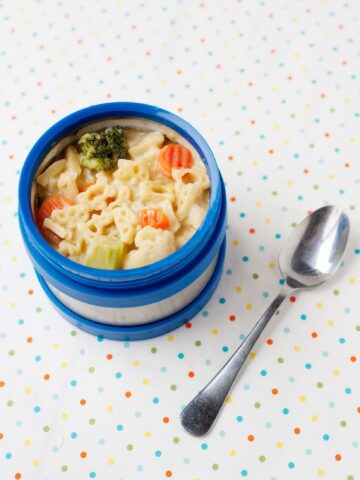 blue thermos with mac and cheese soup with vegetables and a spoon on the side