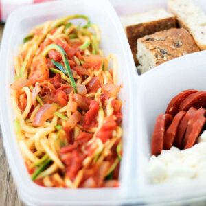 pizza spagetti in a plastic lunch container with cheese, salami and bread slices on the side