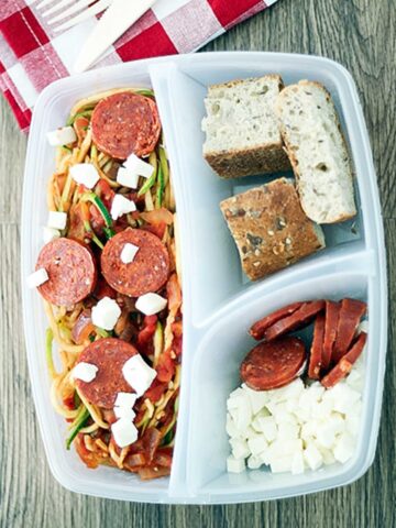 pizza spagetti in a plastic lunch container with cheese, salami and bread slices on the side