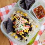 A burrito bowl in a lunchbox made with rice, black beans, corn, cheese, and pico de gallo. Blue corn tortilla chips in the side compartment with salsa in a mini sauce container.