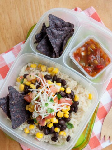 A burrito bowl in a lunchbox made with rice, black beans, corn, cheese, and pico de gallo. Blue corn tortilla chips in the side compartment with salsa in a mini sauce container.