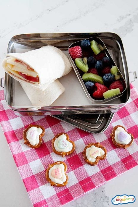 chocolate pretzel hearts on a pink and white cloth napkin next to a lunch container with an apple wrap and sliced blueberries, kiwi, and raspberries