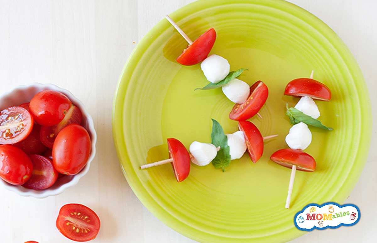 tomato and mozzarella cheese skewers on a green plate with little bowls of tomatoes and cheese on the side. 