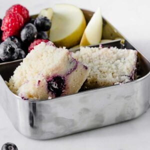 blueberry coffee cake slices in a metal lunchbox with blueberries, strawberries and apple slices on the side