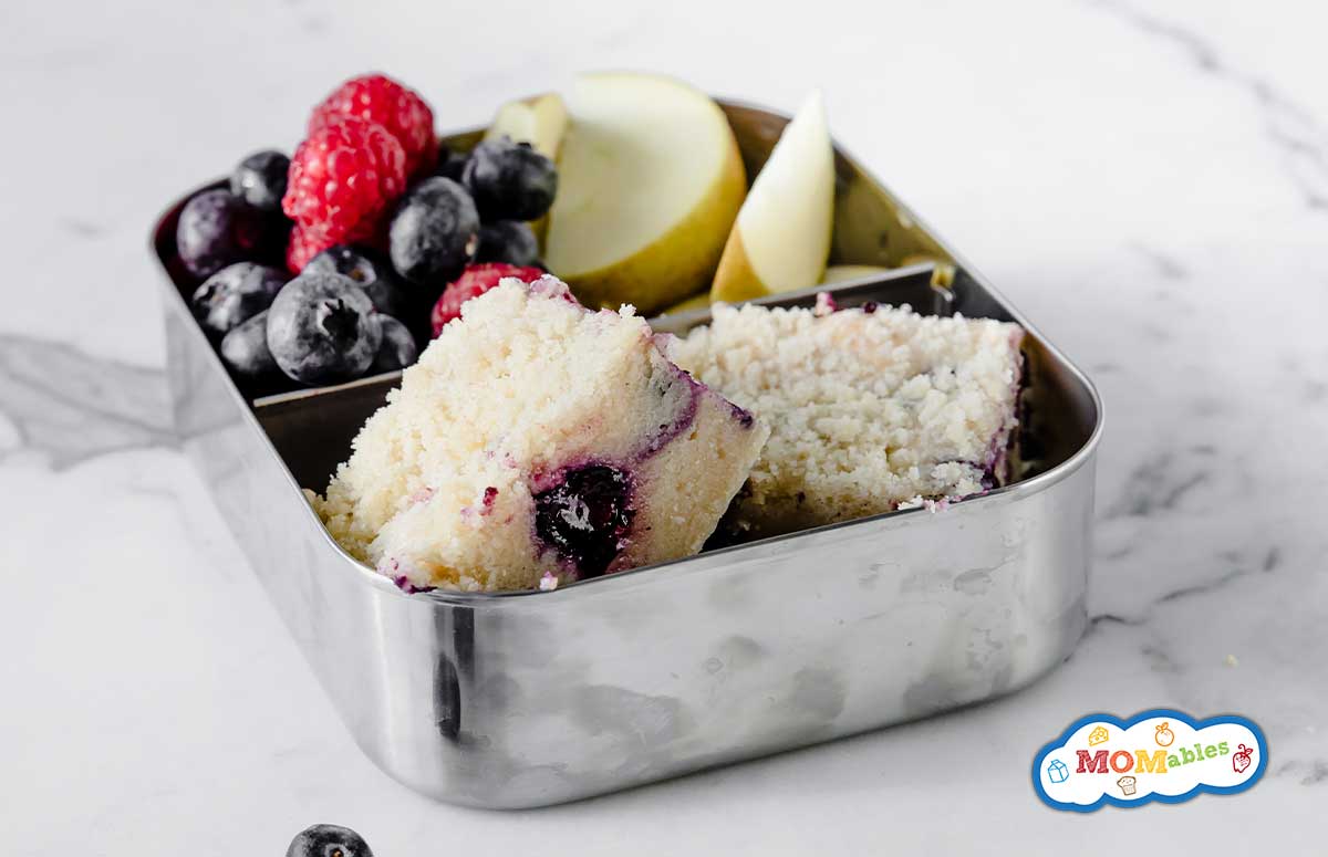 blueberry coffee cake slices in a metal  lunchbox with blueberries, strawberries and apple slices on the side