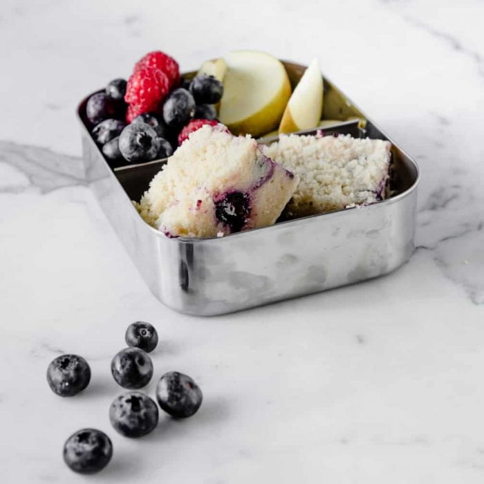 two small squares of blueberry coffee cake in a snack tin with fruit. 