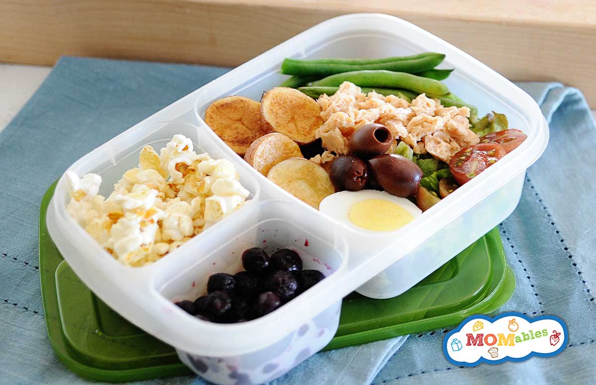 image: salmon salad in a lunchbox with carrots, olives, green beans and tomatoes. Other portions of the lunch container filled with fruit and popcorn.