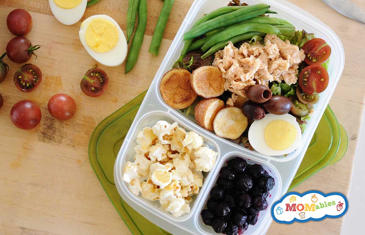 image: salmon salad in a lunchbox with carrots, olives, green beans and tomatoes. Other portions of the lunch container filled with fruit and popcorn.