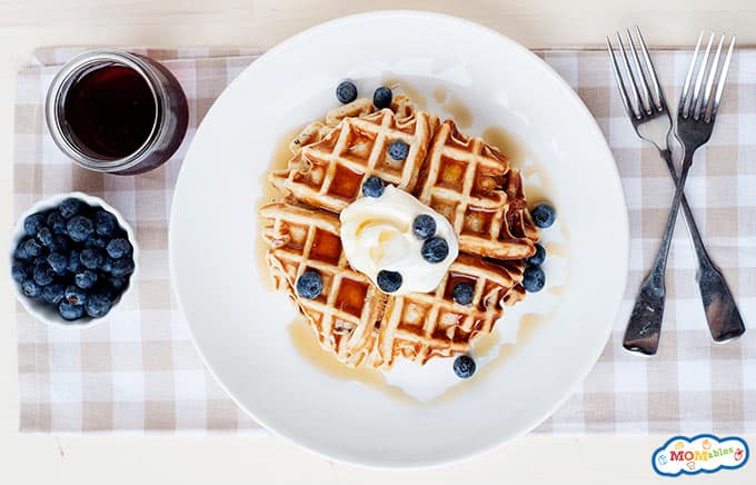 immagine: foto sopra la testa di un waffle su un piatto con due forchette, un piatto di mirtilli e un barattolo di vetro di sciroppo