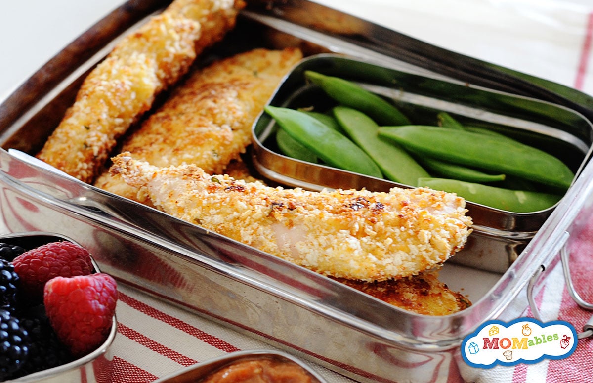 small lunchbox with four baked chicken tenders, and snow peas. Smaller containers hold dip and fresh berries. 