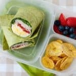 lunchbox with a turkey club wrap in its larger compartment, and banana chips and berries on the smaller ones.