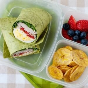 lunchbox with a turkey club wrap in its larger compartment, and banana chips and berries on the smaller ones.