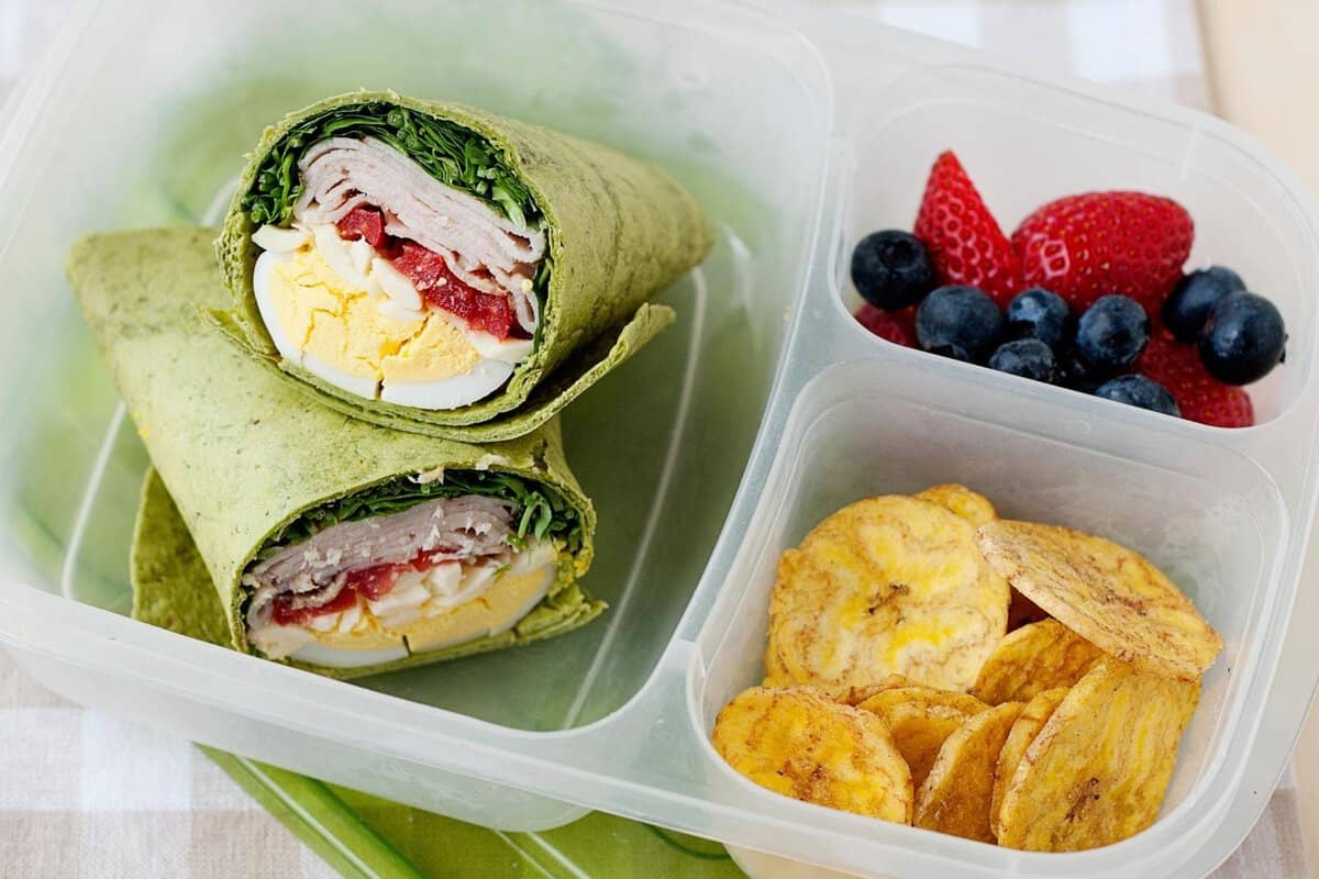 lunchbox with a turkey club wrap in its larger compartment, and banana chips and berries on the smaller ones.