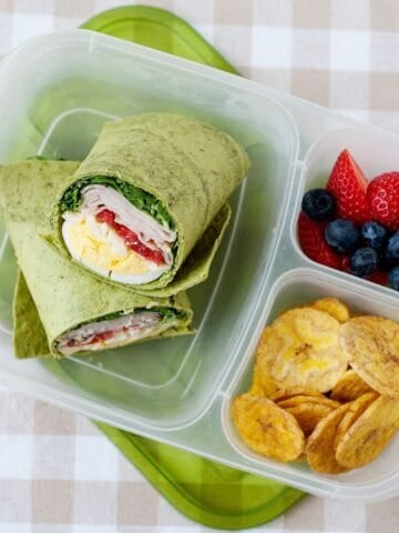 lunchbox with a turkey club wrap in its larger compartment, and banana chips and berries on the smaller ones.