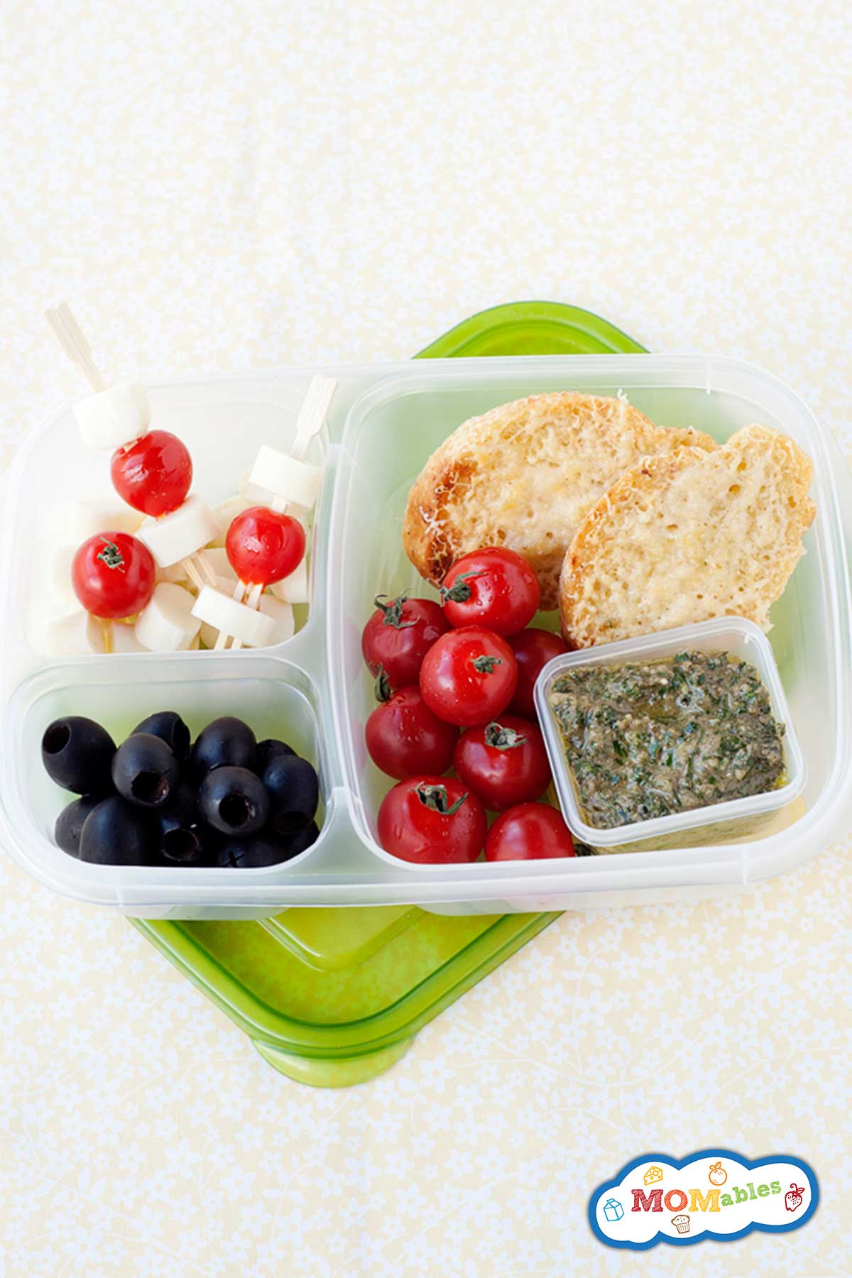 Parmesan crostini, tomato and cheese skewers, and black olives packed into a lunch container with a sauce cup of basil pesto on the side.