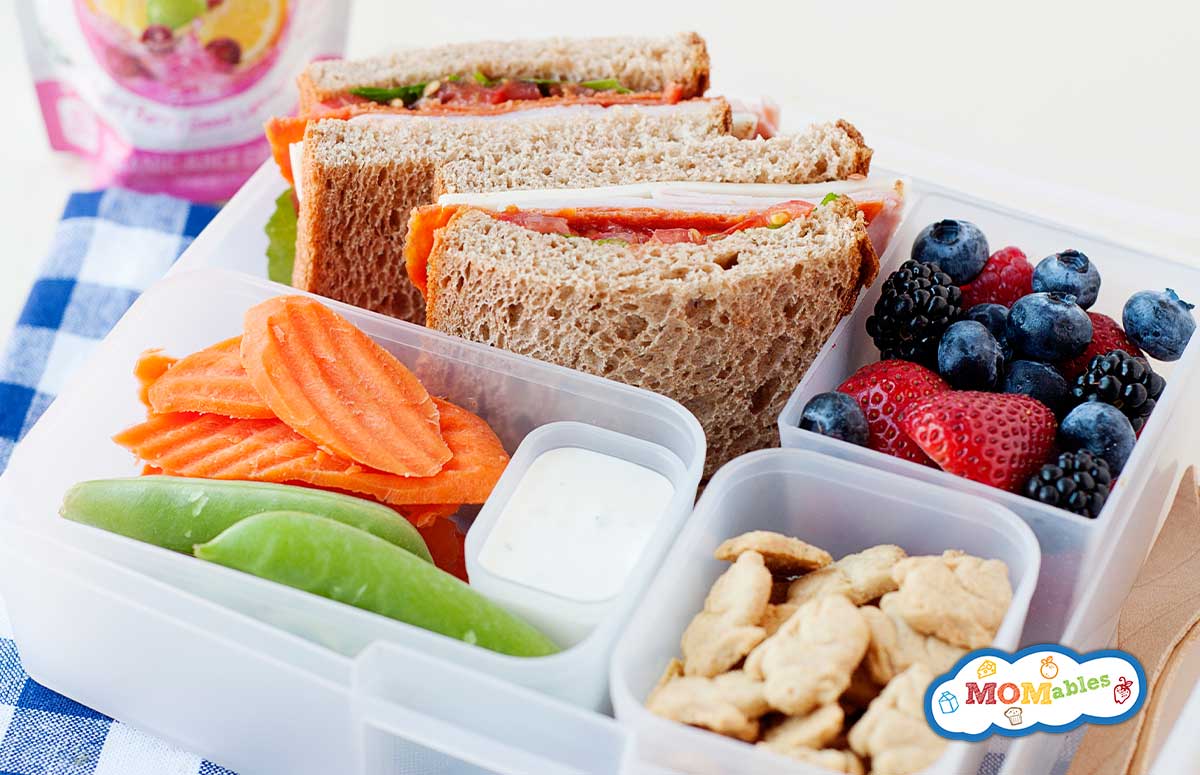 A turkey pepperoni sandwich in a lunchbox with fresh veggies, fruit, animal crackers, and a sauce cup with ranch dressing. 