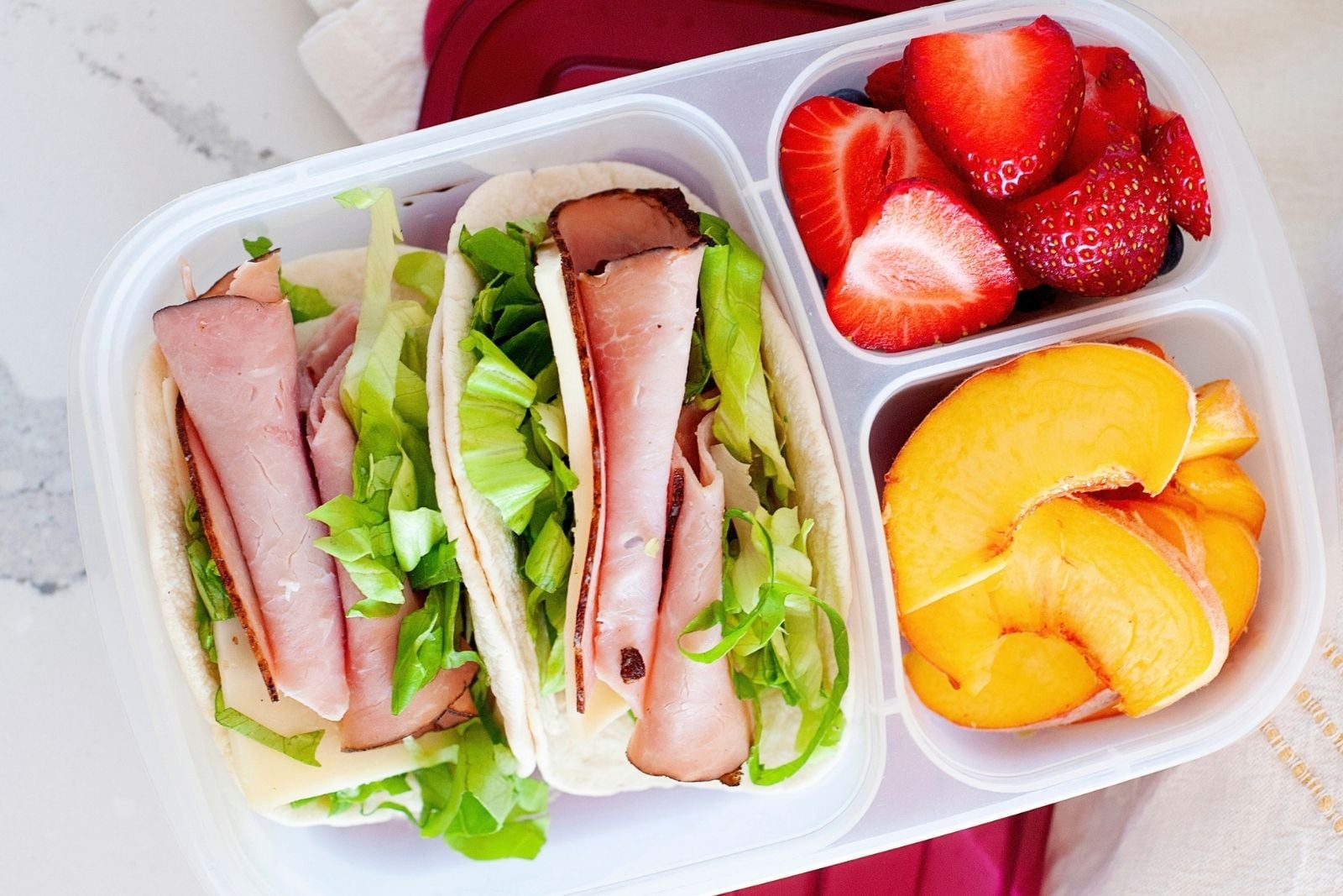 overhead view of a healthy lunch in a compartmentalized container. in the main compartment there are two lunchbox tacos made with ham and cheese. on the side, there are diced strawberries and peaches