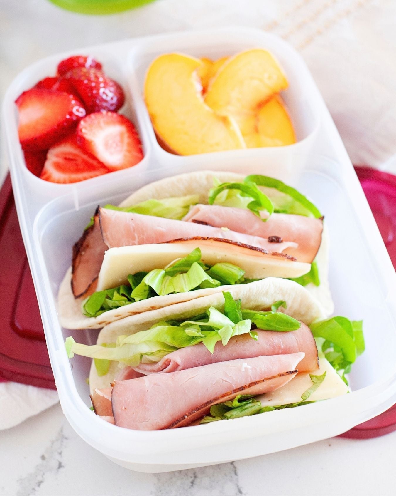 overhead view of a healthy lunch in a compartmentalized container. in the main compartment there are two lunchbox tacos made with ham and cheese. on the side, there are diced strawberries and peaches