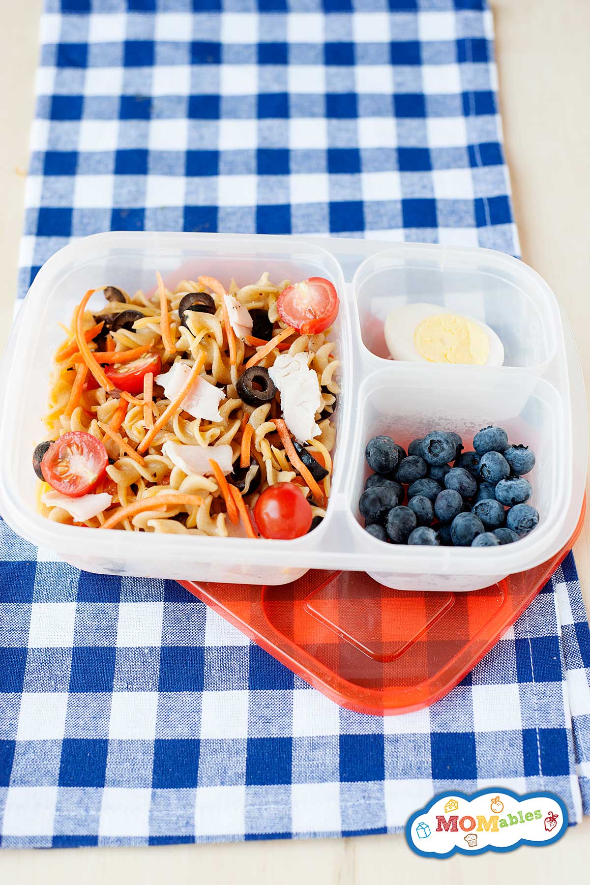 Pasta salad with tomatoes, turkey, carrots, and black olives, packed into a lunch container with blueberries and hard-boiled egg.