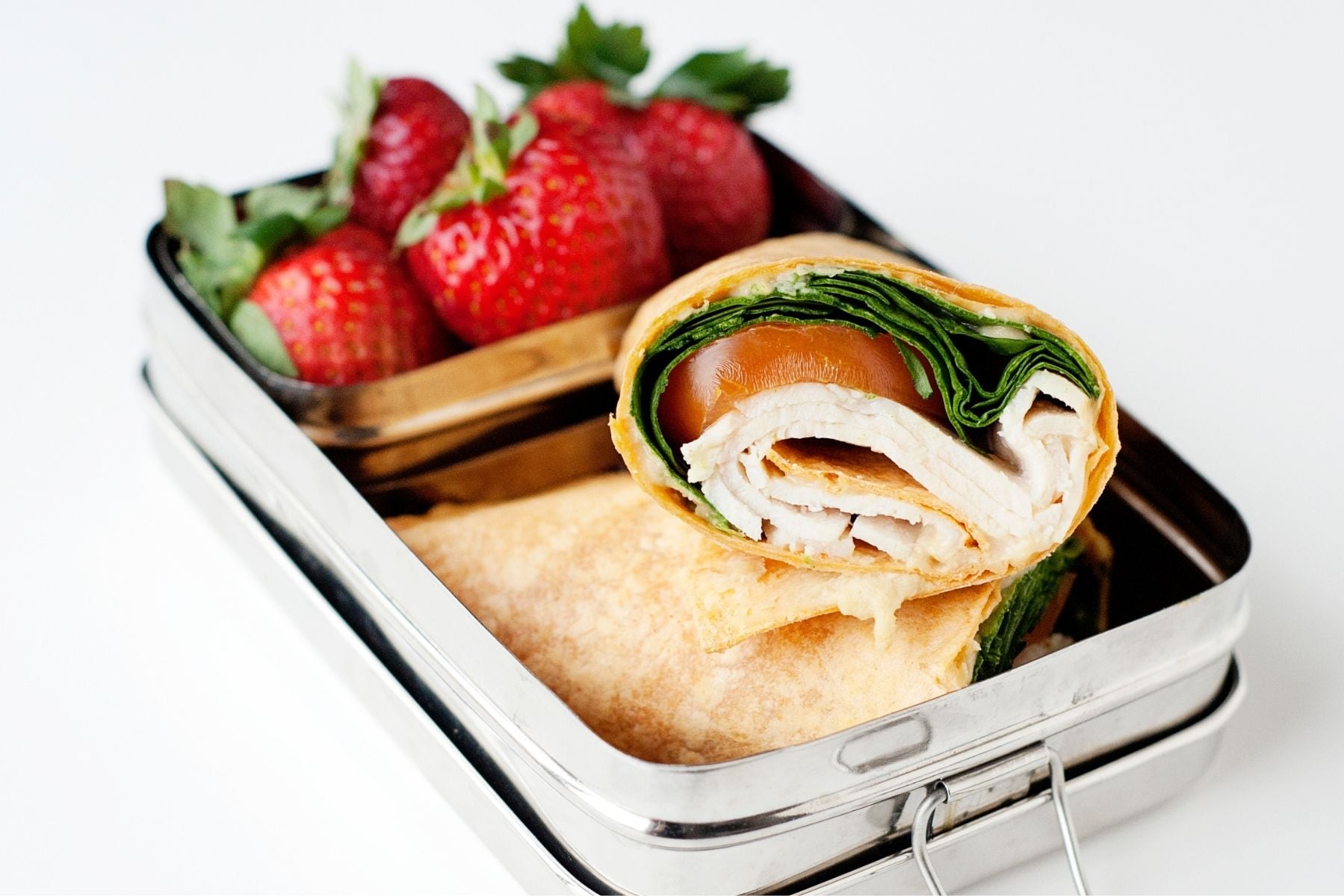 Spinach and turkey wrap sliced in half and packed into a metal lunch container with fresh strawberries