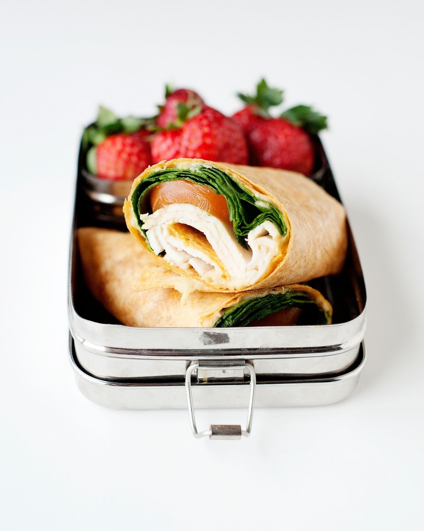 Spinach and turkey wrap sliced in half and packed into a metal lunch container with fresh strawberries.