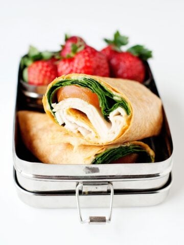 Spinach and turkey wrap sliced in half and packed into a metal lunch container with fresh strawberries.