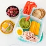 A bento box filled with deli turkey, sliced cheese, hard-boiled egg, red bell pepper, whole grain crackers, and a sauce cup of guacamole with a side container of grapes.