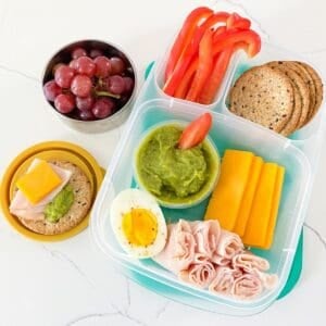 A bento box filled with deli turkey, sliced cheese, hard-boiled egg, red bell pepper, whole grain crackers, and a sauce cup of guacamole with a side container of grapes.