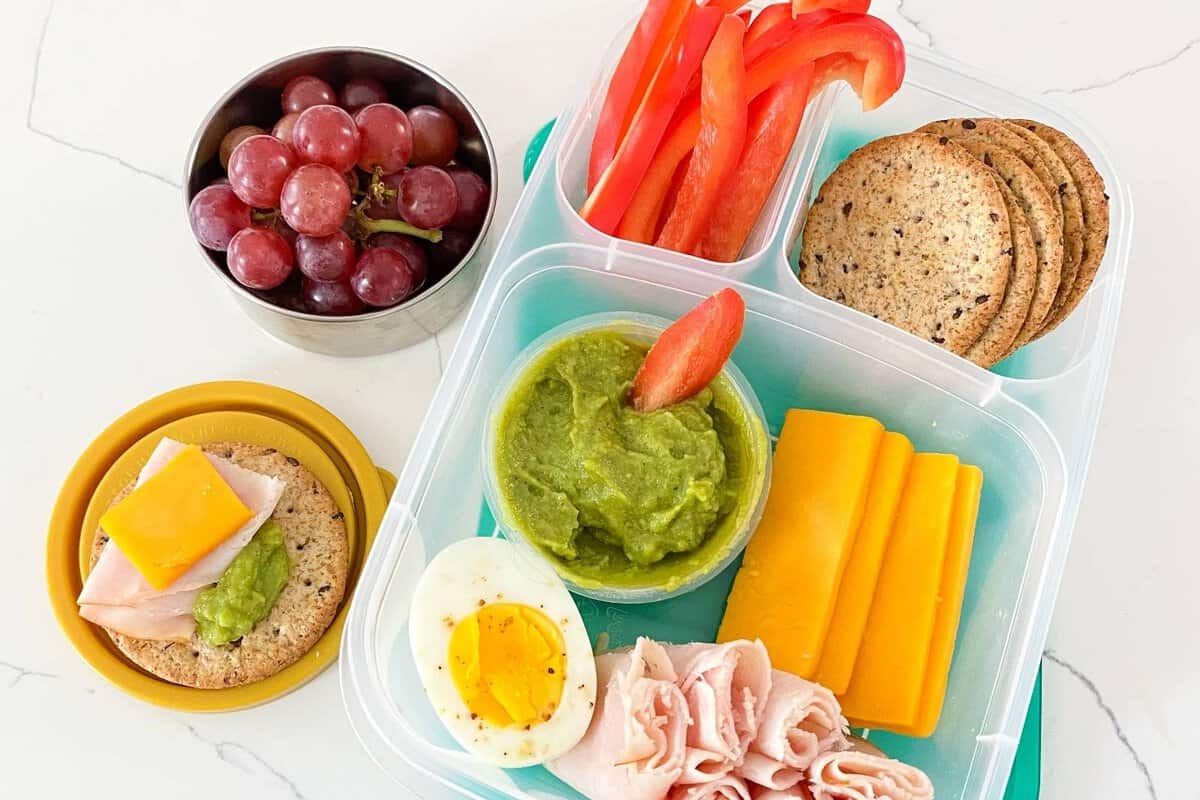 A bento box filled with deli turkey, sliced cheese, hard-boiled egg, red bell pepper, whole grain crackers, and a sauce cup of guacamole with a side container of grapes.