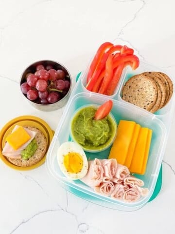 A bento box filled with deli turkey, sliced cheese, hard-boiled egg, red bell pepper, whole grain crackers, and a sauce cup of guacamole with a side container of grapes.