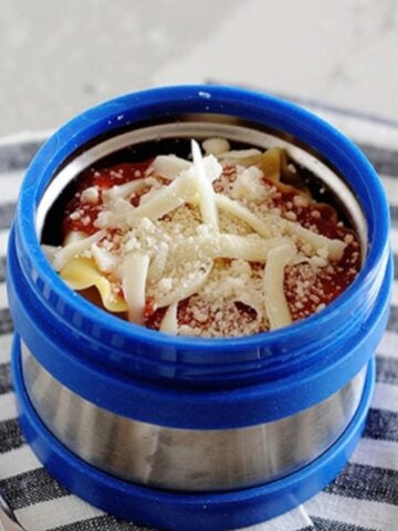 A thermos container filled with lasagna and topped with Mozzarella and Parmesan cheese