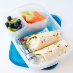 A chicken ranch wrap cut in half and packed into a lunch container with yogurt pack, fresh fruit, and veggies.