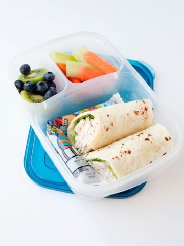 A chicken ranch wrap cut in half and packed into a lunch container with yogurt pack, fresh fruit, and veggies.