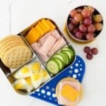 bento box with crackers, hard-boiled eggs, cucumber, turkey and cheese. Another lunchbox with grapes. A cracker with turkey and cheese on the side.
