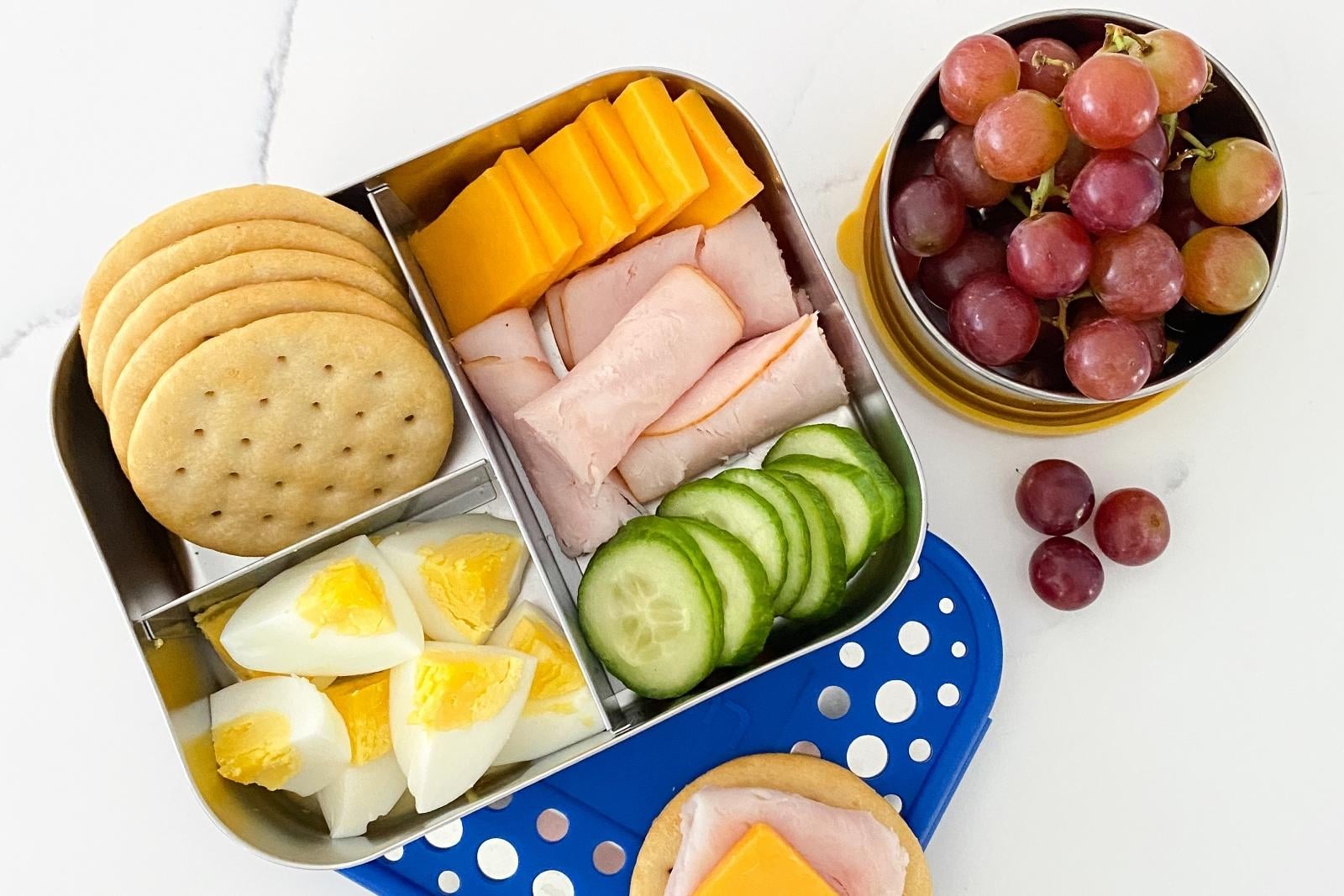 bento box with crackers, hard-boiled eggs, cucumber, turkey and cheese. Another lunchbox with grapes.