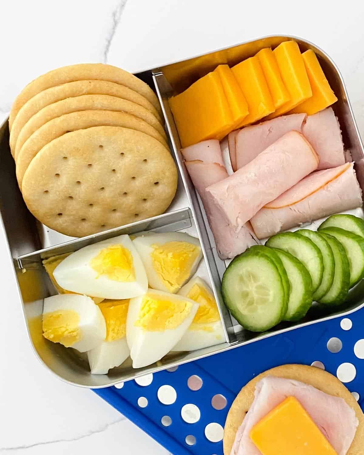 bento box with crackers, hard-boiled eggs, cucumber, turkey and cheese. A cracker with turkey and cheese on the side.