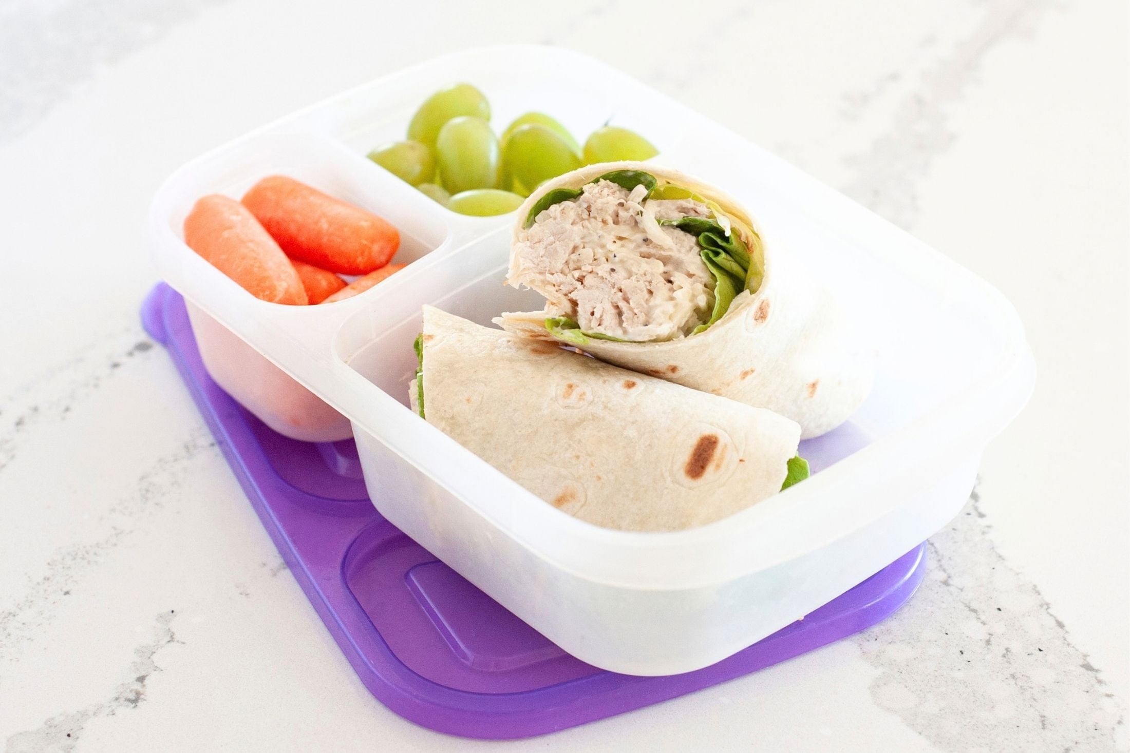 front view of a chicken caesar wrap inside a lunch box, packed with grapes and carrots for a healthy school lunch