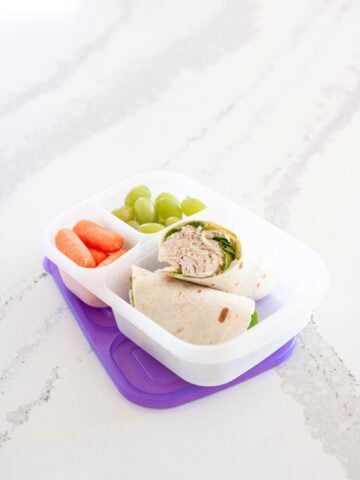 front view of a chicken caesar wrap inside a lunch box, packed with grapes and carrots for a healthy school lunch
