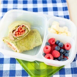 front view of a spinach wrap filled with turkey, hummus, and veggies packed in a lunchbox with berries and cheese in separate compartments