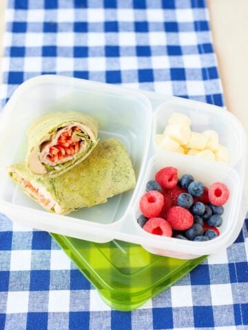 front view of a spinach wrap filled with turkey, hummus, and veggies packed in a lunchbox with berries and cheese in separate compartments