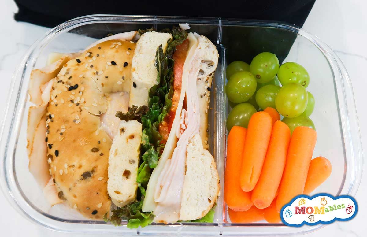 overhead view of a turkey bagel sandwich in a lunchbox with grapes and baby carrots on the side