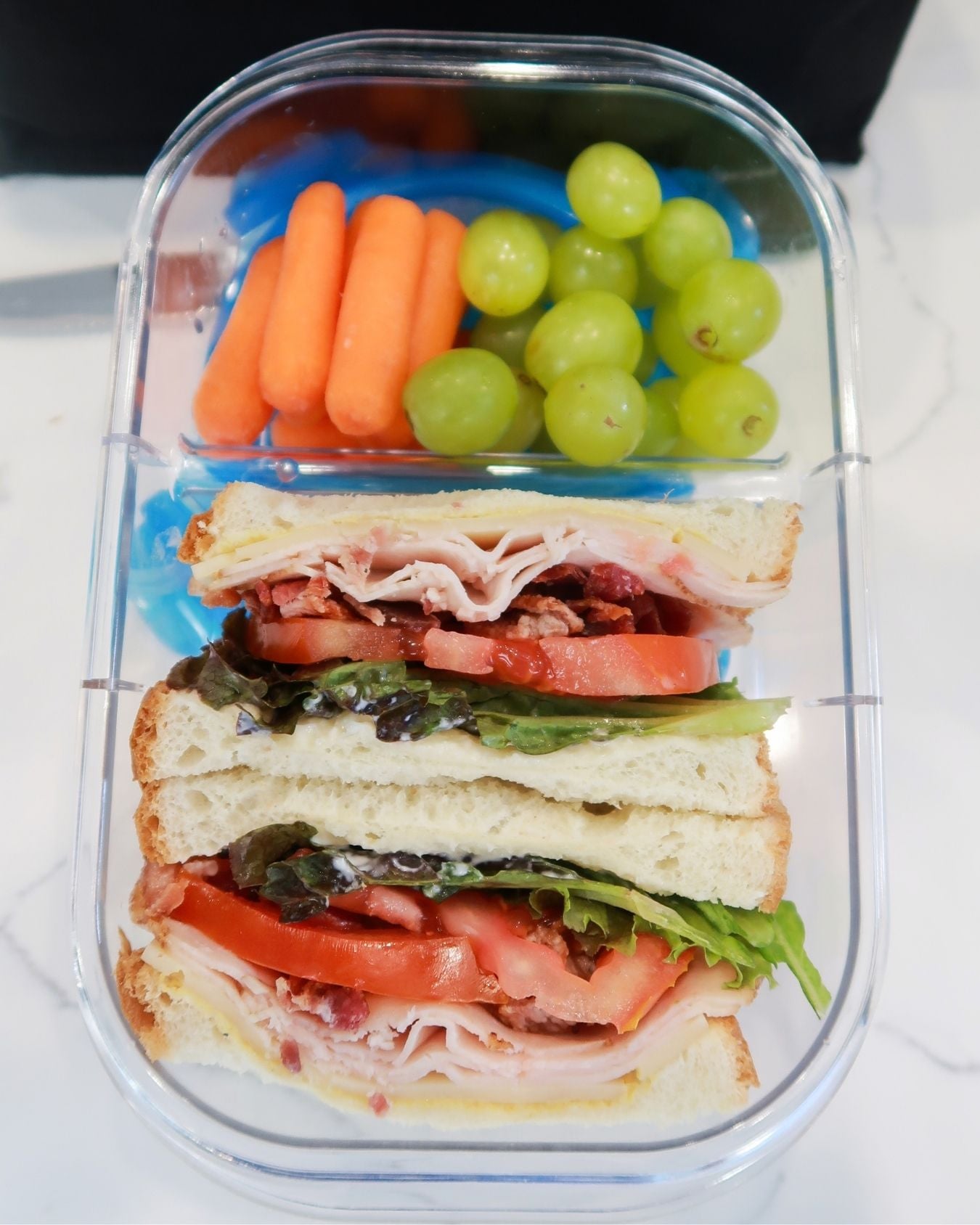 turkey sandwich with bacon, cheddar, tomato and lettuce in a lunchbox with grapes and baby carrots on the side