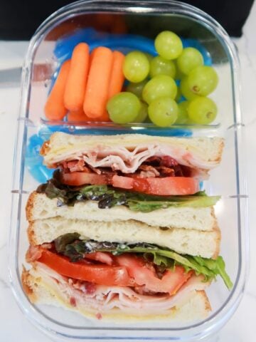 turkey sandwich with bacon, cheddar, tomato and lettuce in a lunchbox with grapes and baby carrots on the side