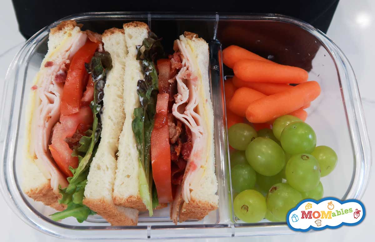 turkey sandwich with bacon, cheddar, tomato and lettuce in a lunchbox with grapes and baby carrots on the side