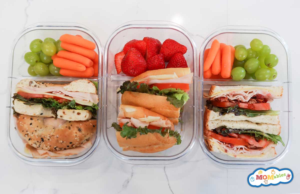 three turkey sandwiches next to each other in lunchoxes: a turkey bagel sandwich, a turkey sub sandrich and a turkey and bagel sandwich with grapes, strawberries and baby carrots on the side